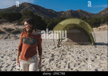 Attraktive junge Frau, die während eines Campings an einem windigen und sonnigen Frühlingstag am Strand spazieren geht. Zelt im Hintergrund. Stockfoto