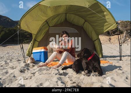 Junge attraktive Frau, die auf dem Sand sitzt und ein Tablet hält, während sie lächelt und den Hund anschaut, der neben ihr liegt. Camping Zelt am Strand. Stockfoto