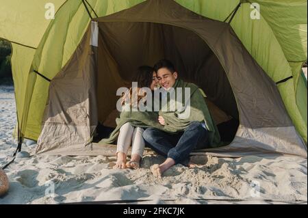 Junges Paar, das im Campingzelt sitzt und mit lächelnden Gesichtern nach draußen blickt, umarmt und eine Decke trägt. Stockfoto