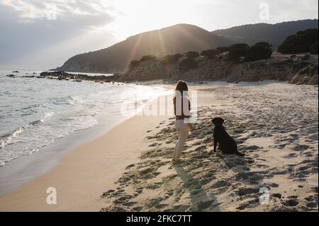 Rückansicht Frau, die am Strand steht und anschaut Der Horizont mit ihrem Hund, der in die gleiche Richtung schaut Stockfoto
