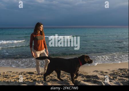 Frau, die am Strand steht und den Horizont mit ihrem Hund in die gleiche Richtung blickt. Stockfoto