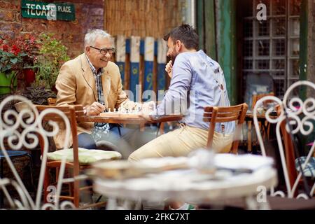 Ein älterer Mann und sein junger Freund haben eine gute Zeit, während sie in einer fröhlichen Atmosphäre in der Bar ein Schach spielen. Freizeit, Bar, Freundschaft, Outdoor Stockfoto