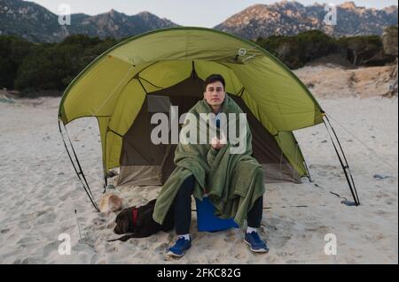 Junger Mann, der eine Decke trägt, um sich während eines Campingzelts am Strand mit seinem Hund aufzuwärmen. Stockfoto