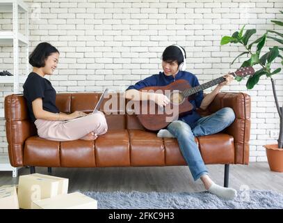 Junge Liebhaber verbringen Zeit zusammen auf Urlaub im Wohnzimmer. Asiatischer Mann mit Kopfhörern spielt eine akustische Gitarre, während das Mädchen das Paket überprüfen Stockfoto