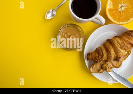 Frühstück: Croissant mit Erdnusspaste, schwarzem Kaffee und Orange auf dem gelben Hintergrund.Draufsicht.Copy space. Stockfoto