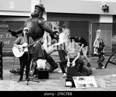 Straßenmusiker unterhalten Passanten vor der verzweifelten Dan-Statue in der High Street, Dundee, während Minnie, der Minx, auf sie schaut. Stockfoto