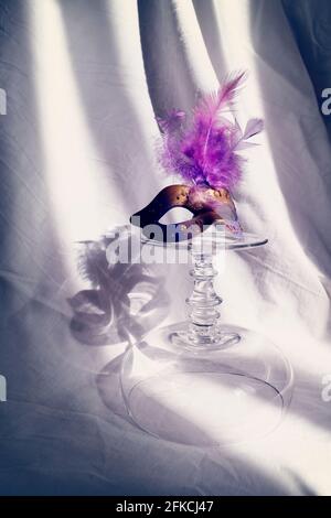Ein umgedrehtes Champaign-Glas und eine Maskerade darauf. Gatsby-Vibes. Schöne Schatten im Hintergrund geben Rätsel auf Stockfoto
