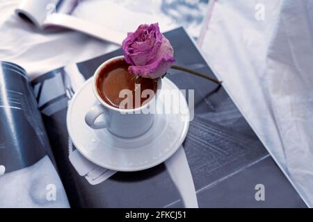Ein türkischer Kaffeebecher und eine verdirte Rose auf dem Becher. Einige offene Zeitschriften darunter. Einige weiße Laken darunter. Romantische Morgenroutine. Stockfoto
