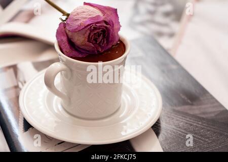 Türkischer Kaffee mit Rosengeschmack. Ein türkischer Kaffeebecher und eine getrocknete Rose auf der Oberseite des Getränks. Einige offene Zeitschriften darunter. Stockfoto