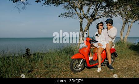 Liebespaar auf rotem Motorrad in weißer Kleidung auf Waldweg-Tour. Zwei kaukasische Touristen Frau und Mann küssen Umarmungen sitzen auf dem Roller. Stockfoto