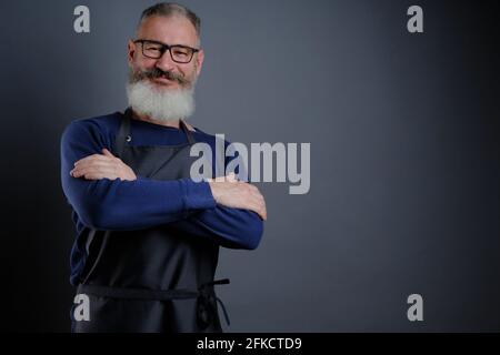 Porträt eines hübschen, reifen bärtigen, auf grauem Hintergrund isolierten Mannes mit Schürze, kaukasischer Arbeiter mit lächelndem Bart Stockfoto