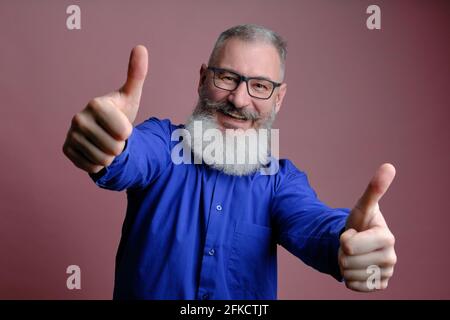 Porträt eines reifen bärtigen Mannes mit blauem Hemd und Daumen nach oben auf rosa Hintergrund, kaukasischer Mann mit lächelndem Bart Stockfoto