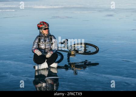 Frau, die in der Nähe ihres Fahrrads auf Eis sitzt. Die Radfahrerin hörte auf zu ruhen. Sie sitzt auf dem Rad und genießt die schöne Aussicht auf den Sonnenuntergang. Stockfoto