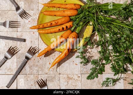 Ein paar gegrillte Karottenwurzeln mit grünen Blättern und Salz Flocken auf einem grünen Bastelteller auf Steingeschichten Erdtöne Hintergrund mit sechs Gabeln oben vie Stockfoto