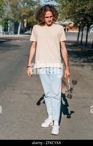 Vertikales Porträt eines attraktiven rothaarigen jungen Mannes, der auf der Straße läuft. Er hat lange lockige Haare. Er hält ein Skateboard und blickt auf Prot hinunter Stockfoto