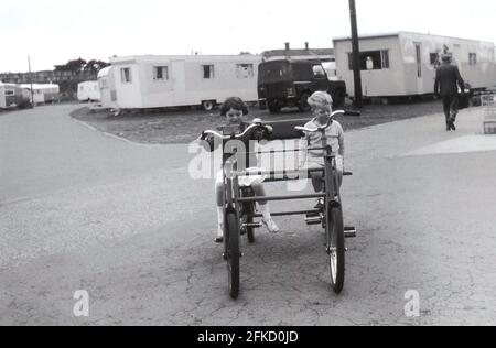 1964, historisch, draußen auf einem Pfad in einem Ferienpark, sitzt ein kleiner Junge mit seiner kleinen Schwester auf einem großen, metallumrahmten „Feriencamp“-Typ mit zwei oder zwei Sitzern, Suffolk, England, Großbritannien. Diese robusten Doppelsitzer-Fahrräder waren ein großer Spaß und eine gute Möglichkeit, sich auf den breiten Wegen auf dem Gelände zu bewegen. Stockfoto