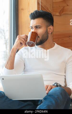 Indischer Mann nippte Kaffee und arbeitete an seinem Laptop. Sitzen neben dem Fenster in einem ruhigen, gemütlichen Zimmer. Trägt ein weißes Hemd und Jeans. Wintersaison in der Stadt Zakopane, Polen. Stockfoto