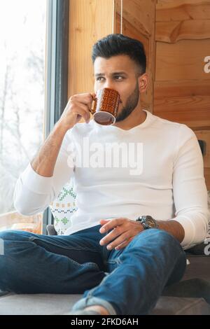 Indischer Mann, der sich zu Hause in einem gemütlichen Zimmer entspannt. Schlürfen Sie Kaffee neben dem Fenster und schauen Sie nach draußen durch das Glas des Fensters. Wintersaison in der Stadt Zakopane, Polen. Stockfoto