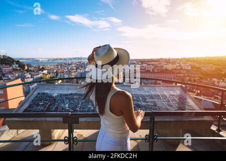 Junge, unbeschwerte Touristin in weißem Hut, die Lissabon aus der Sicht beim Sonnenuntergang betrachtet. Tourismus in Europa. Reise-, Urlaubs- und Urlaubskonzept. Stockfoto