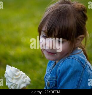 Nahaufnahme eines niedlichen, braunhaarigen, blauäugigen Mädchens, das an einem sonnigen Tag eine blaue Denim-Jacke trägt und ein Eis im Gras isst Stockfoto