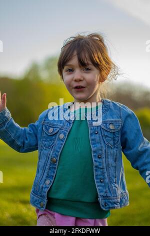 Porträt eines niedlichen, braunhaarigen, blauäugigen Mädchens, das an einem sonnigen Tag in einem Park läuft und eine blaue Jacke und einen grünen Pullover trägt Stockfoto