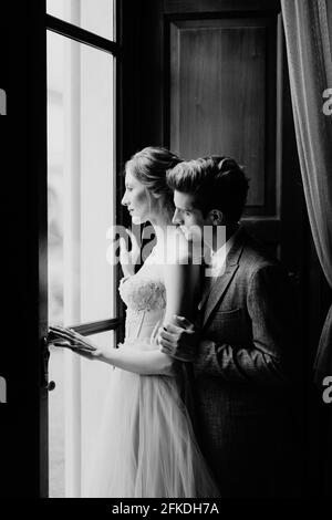 Der Bräutigam umarmt die Braut von hinten am großen Fenster einer alten Villa. Comer See. Schwarzweiß-Foto Stockfoto