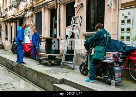 Zwei Männer, die Kabel verlegen, stellen ein Problem für die Frau dar, die die Post in einer Gasse in der Nähe von Huai Hai Zhonglu im Zentrum von Shanghai, China, liefert. Stockfoto