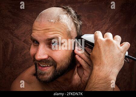 Kaukasischer Mann, der versucht, seinen Kopf mit einem elektrischen Rasiermesser zu rasieren. Ein brutaler Glatze hält einen Rasierer in der Hand und rasiert Stoppeln auf einem metallischen Hintergrund Stockfoto