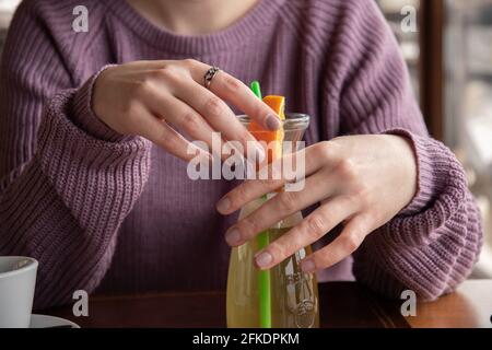 Weibliche Hände halten Flasche mit Trinkhalm von frischem Detox-Zitrussaft. Nahaufziehfinger einer jungen Frau mit violettem Strickpullover Stockfoto