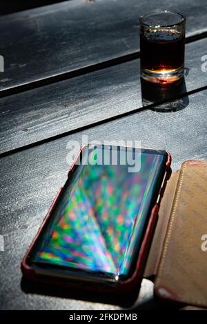 Lebendige Regenbogenstrahlen spiegeln sich auf dem Handy-Bildschirm auf dem schwarzen Tisch mit einem Glas mit Alkoholgetränken wider. Defokussierte mehrfarbige Strahlen und Reflexion in Smart Stockfoto