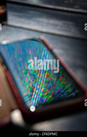 Helle Regenbogenstrahlen spiegeln sich auf der Bildschirmoberfläche des Mobiltelefons mit dunklem, unscharfem Hintergrund wider. Mehrfarbige Strahlen und Reflexion in Smartphone-Bildschirm closeu Stockfoto
