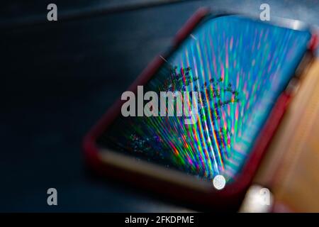 Helle Regenbogenstrahlen spiegeln sich auf der Bildschirmoberfläche des Mobiltelefons mit dunklem, unscharfem Hintergrund wider. Mehrfarbige Strahlen und Reflexion in Smartphone-Bildschirm closeu Stockfoto