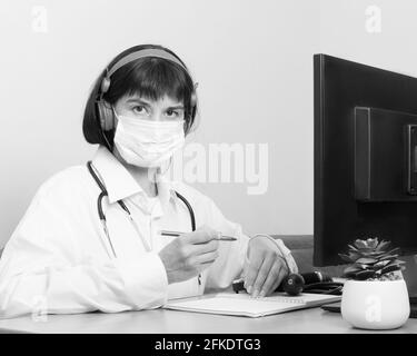 Eine Ärztin in einer medizinischen Schutzmaske schaut Sie an, während sie an einem Tisch in einem Büro sitzt, schwarz und weiß. Der Arzt hört dem Patienten zu Stockfoto