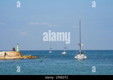 Jaffa, Israel - 31. März 2021: Drei Boote, die an einem sonnigen Tag in den Hafen von Jaffa, Israel, fahren, an einem Leuchtturm vorbei. Stockfoto