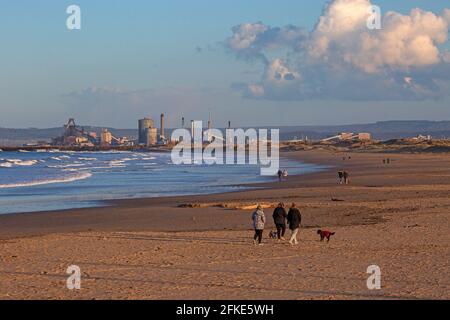 Leute, die am Strand entlang laufen, mit den Redcar Stahlwerken in der Ferne, England, Großbritannien Stockfoto