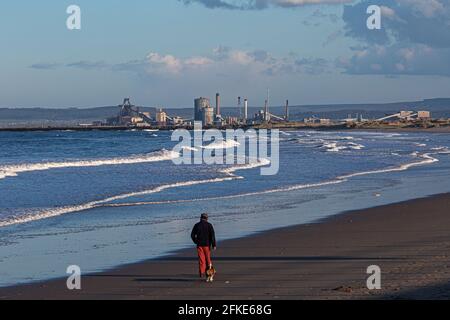 Mann und sein alter Hund laufen am Strand entlang mit dem Redcar Stahlwerk in der Ferne, England, Großbritannien Stockfoto