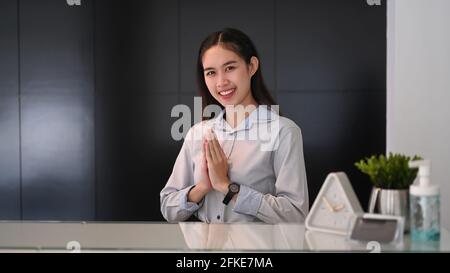 Die junge asiatische Rezeptionistin, die an der Rezeption sitzt und die Hände hebt, respektiert die Gäste im Hotel. Stockfoto