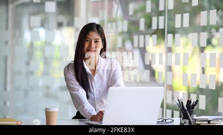 Fröhliche junge Angestellte, die im modernen Büro mit einem Laptop sitzt und vor der Kamera lächelt. Stockfoto