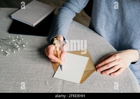 Weibliche Hände schreiben Hochzeitseinladungskarte. Unbeschriftete Papierkarte, Umschlag, Notizbuch, Gypsophila auf Tisch mit Leinentischdecke. Flach liegend, Draufsicht Stockfoto
