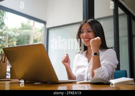 Glückliche, erfolgreiche Geschäftsfrau, die die Arme hochhob, weil sie den Erfolg des Start-ups feierte und sich einen Laptop ansah. Stockfoto