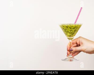 Frau Hand hält Kiwi Obst Smoothie Shake mit Stroh in der martini-Glas gegen beige Wand. Stockfoto