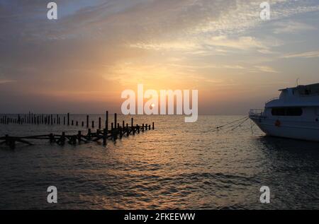 Sonnenaufgang auf dem Meer. Hölzerne Stapel zerstörter Pier ragen aus dem Wasser und dem Boot. Morgensonne. Rotes Meer, Safaga, Ägypten. Stockfoto