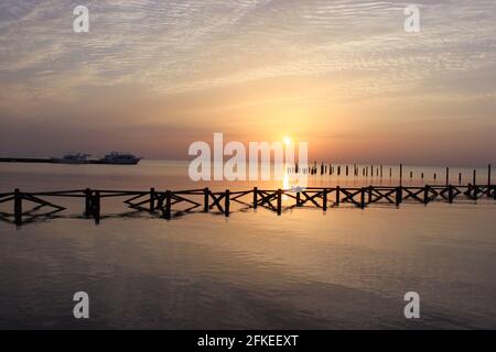 Goldener Sonnenaufgang auf dem Meer. Holzstapel von alten zerstörten Pier ragen aus dem Wasser. Das Schiff fährt in der Ferne von der Marina ab. Morgensonne Stockfoto