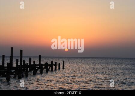 Sonnenaufgang auf dem Meer. Holzstapel von zerstörten Pier ragen aus dem Wasser. Die Sonne guckt hinter den Wolken hervor. Rotes Meer, Safaga, Ägypten. Stockfoto