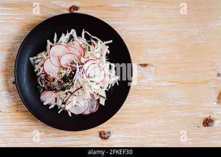 Schüssel mit frischem Salat auf rustikalem Holzhintergrund. Koleslaw mit Rettich auf schwarzem Teller von oben gesehen, gesunde vegane Mahlzeit Stockfoto