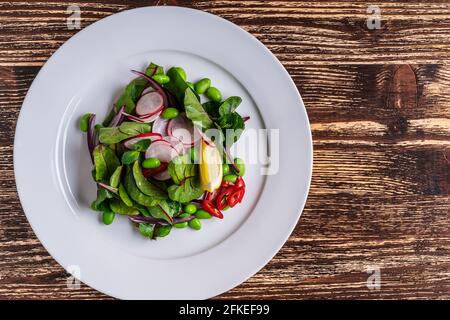 Schale mit frischem Frühlingssalat auf rustikalem Holzhintergrund. Mangold Blätter mit Rettich auf weißem Teller von oben gesehen, gesunde vegane Mahlzeit. Stockfoto