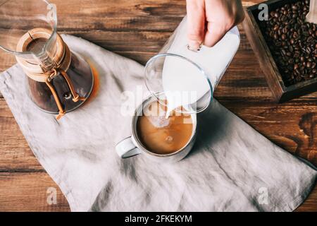 Gießen Sie Milch mit einer Glaskaraffe in eine Tasse mit Kaffee auf einem braunen Holztisch Stockfoto