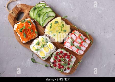 Sechs Sandwiches mit frischen Karotten, Gurken, Ananas, roten Johannisbeeren, Krabbenstäbchen und Wachteleiern mit Erbsen-Mikrogemüse auf Holzbrett auf grauem Backgro Stockfoto