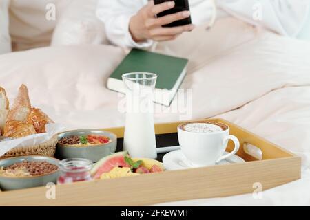 Tasse Kaffee, ein Glas Joghurt, Obst, Brot und Müslischale auf einem Holztablett vor der Frau, die im Bett liegt Stockfoto