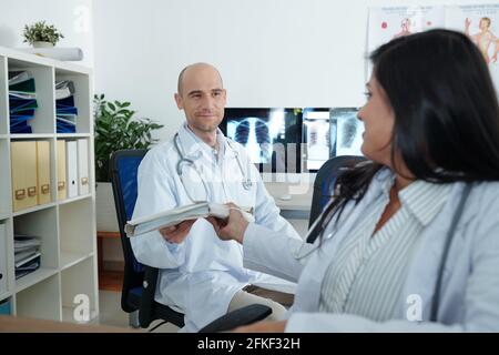 Der Arzt gibt ihrem lächelnden Kollegen einen Ordner mit Dokumenten Sie arbeiten mit Papieren im medizinischen Büro Stockfoto